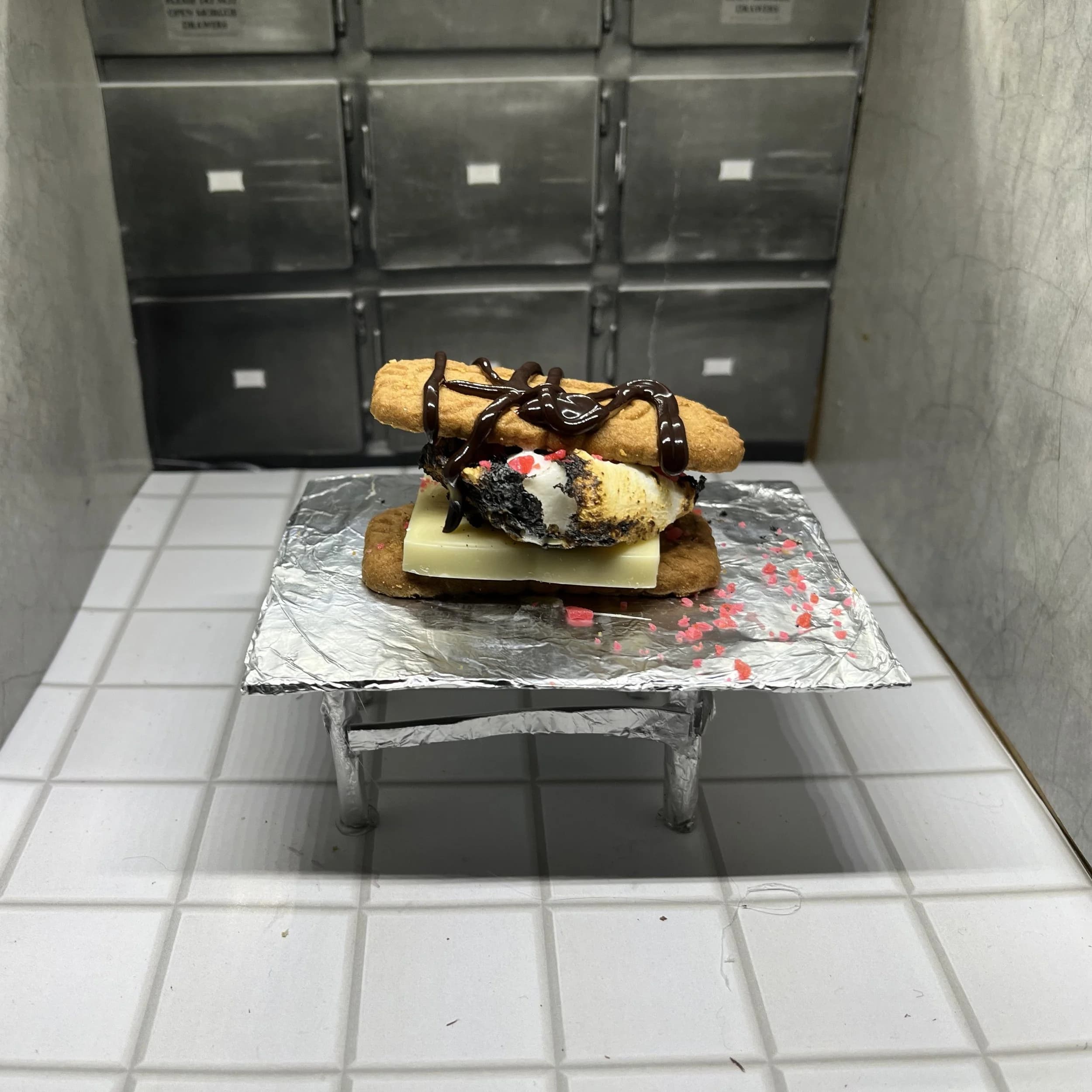A s'more on a foil tray inside a miniature morgue drawer set
