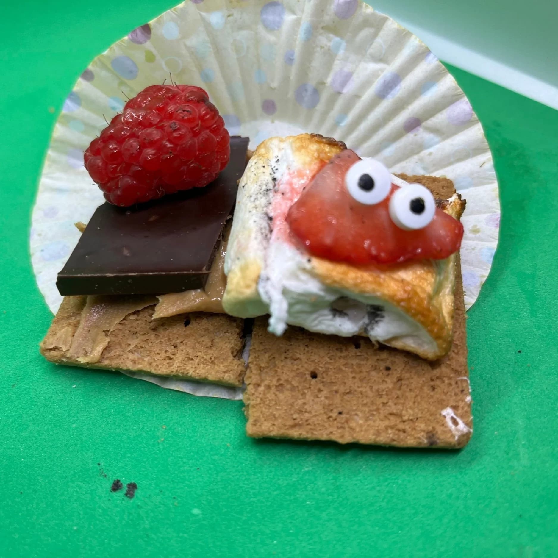 A s'more topped with a raspberry, dark chocolate and a marshmallow with candy eyes, on a graham cracker